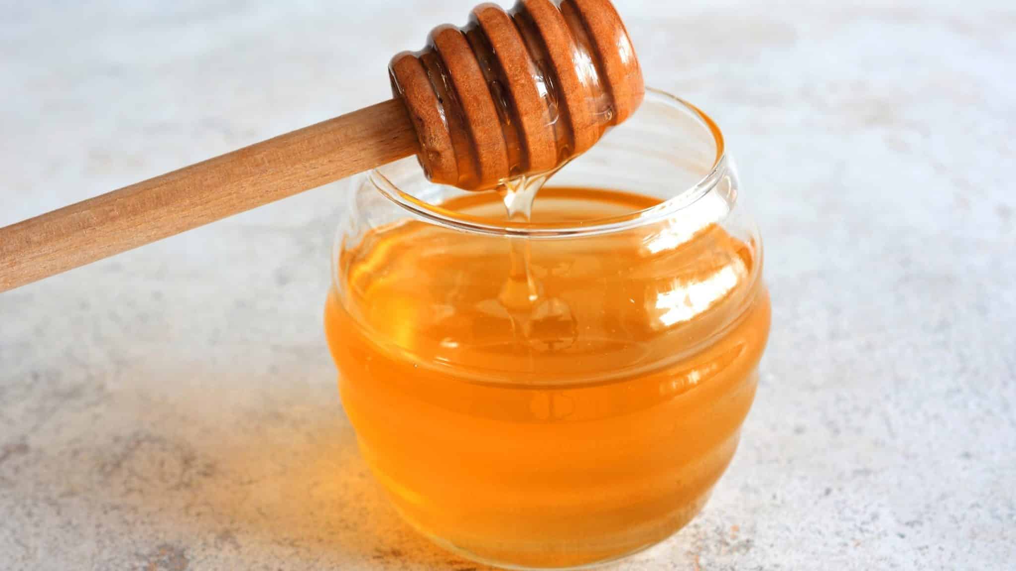 A wooden honey dipper dripping honey into a glass jar of honey