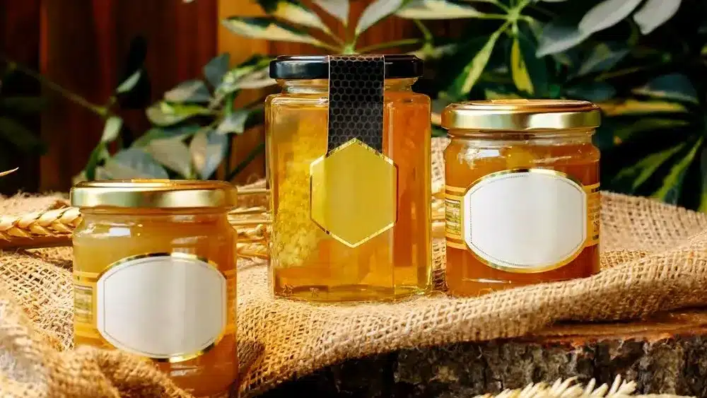 Three jars of honey with plain labels, set on burlap fabric with greenery in the background